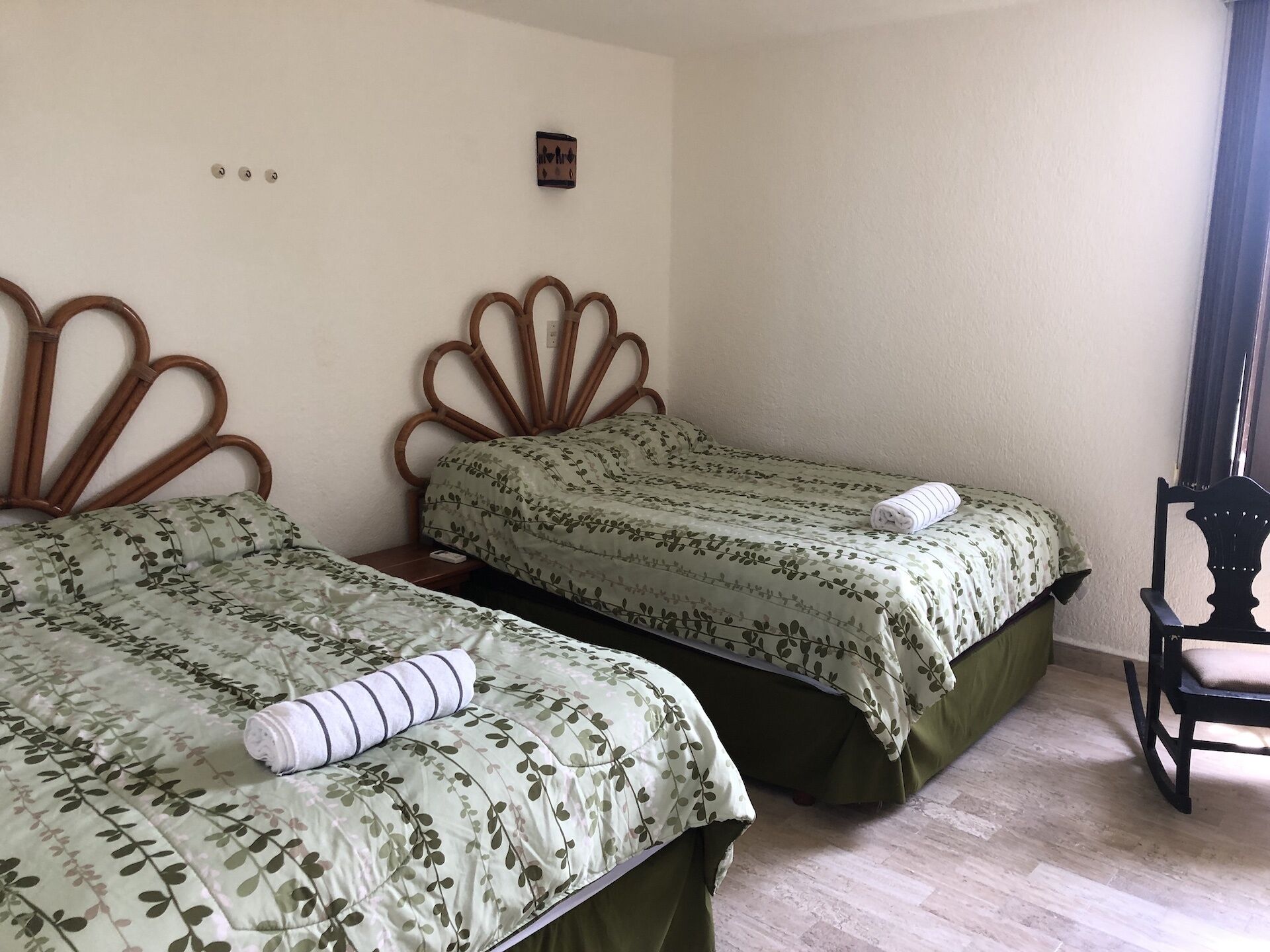 A room with two beds, each with a wooden headboard and green floral bedding. There is a small table with a lamp and a chair.