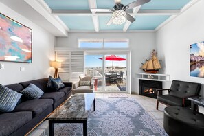 A spacious living room with a dark sofa, a coffee table, and a fireplace. There is a large window with a view of the outdoors, and a ceiling fan. - Ocean Club - Oceanfront 4 BR (Ocean City)