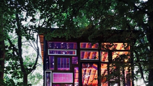 A multi-level wooden structure with illuminated windows, a wooden deck, and a seating area with two people. - Wall of Windows Treehouse-Romantic Woodland Retreat-Escape (Doyle)