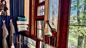 A bathroom with a bathtub, a wooden chair, a guitar leaning against the window, and a view of trees outside. - Wall of Windows Treehouse-Romantic Woodland Retreat-Escape (Doyle)