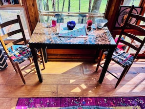 A wooden table with a board game, a blue cap, and a candle holder. Two wooden chairs with colorful cushions are positioned around the table. - Wall of Windows Treehouse-Romantic Woodland Retreat-Escape (Doyle)