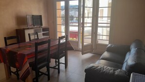 A living room with a dining table, chairs, a sofa, a TV on a cabinet, and a sliding glass door leading to a balcony. - 1 Bedroom Apartment in Cassis (Cassis)