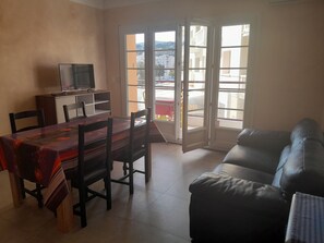 A living room with a dining table, chairs, a sofa, a TV on a cabinet, and a sliding glass door leading to a balcony.