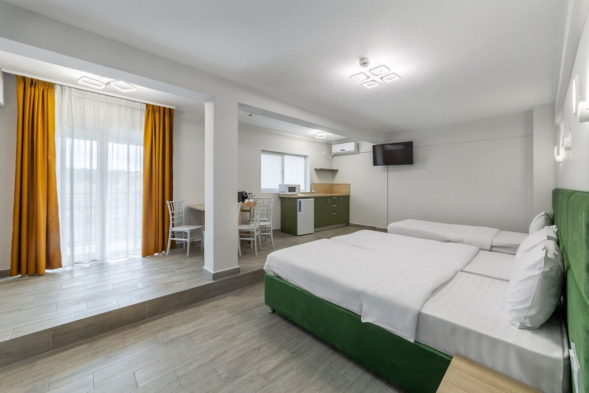 Habitación de hotel con cama verde, cortinas blancas, escritorio con sillas, kitchenette y televisor en la pared.