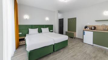 A modern hotel room with a green bed, white bedding, a small kitchenette, and a separate bathroom.