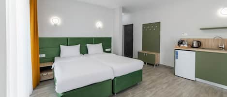 A modern hotel room with a green bed, white bedding, a small kitchenette, and a separate bathroom.