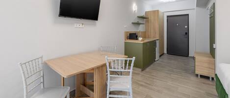 Quarto compacto com mesa de jantar de madeira, duas cadeiras brancas, televisão de tela plana na parede, cozinha compacta com micro-ondas e uma porta que dá para outro quarto.