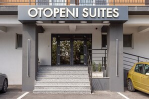 La entrada a Otopeni Suites, con una escalera que lleva a la puerta, un coche amarillo aparcado a la derecha y una fachada de edificio moderna.