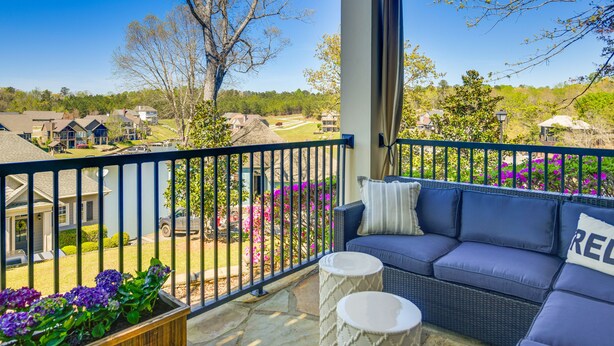 Un balcon avec un canapé en osier, des petites tables rondes blanches et une vue sur un quartier résidentiel avec des maisons et des arbres.