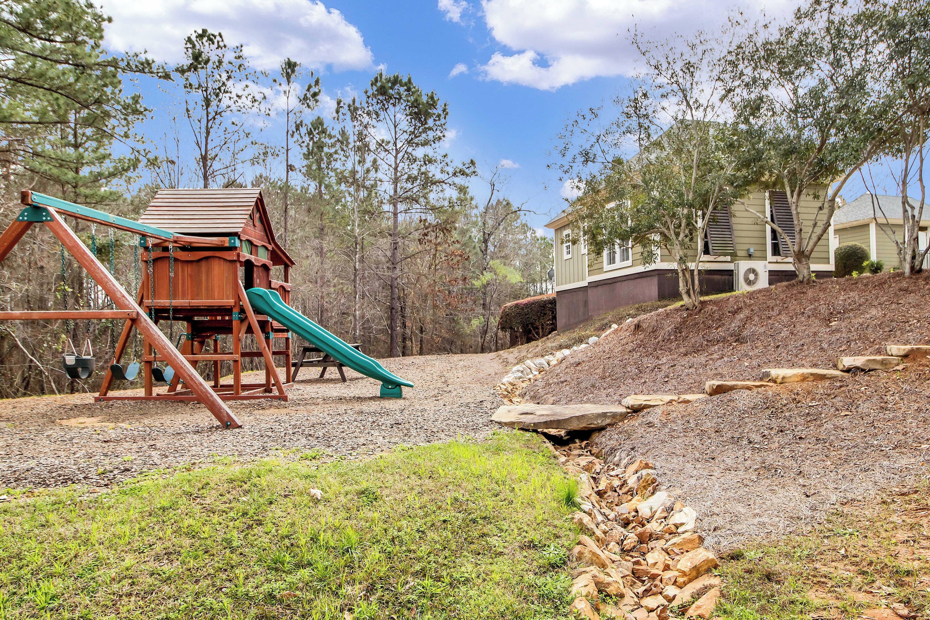 A playground with a swing set and slide, surrounded by trees and a house in the background.