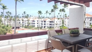 A balcony with wicker furniture, a potted plant, and a view of a resort with palm trees and a swimming pool.