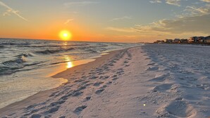 Uma praia com pegadas que levam à água, um pôr do sol ao fundo e edifícios ao longo da costa.