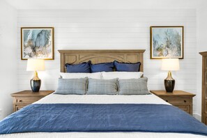 Una cama bien hecha con colcha azul, dos mesitas de noche con lámparas, y dos cuadros abstractos enmarcados en la pared.