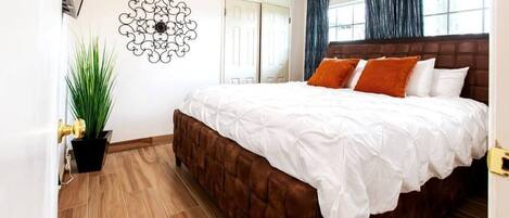 A bedroom with a large bed, a potted plant, a decorative wall piece, and a door.
