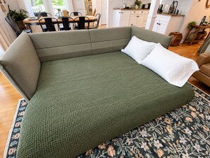 A green sofa with a white pillow in a room with a wooden floor, a dining table, and a kitchen area in the background.