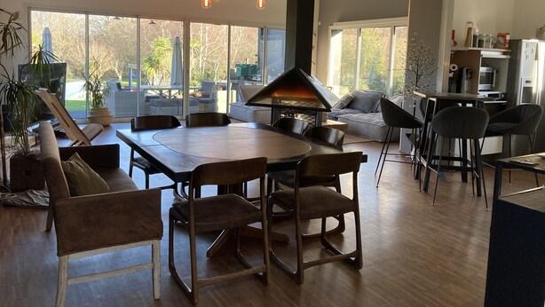 A spacious living room with a large dining table, multiple chairs, a fireplace, and large windows offering a view of the outdoors. - Villa 290m2, Piano, Piscine et Spa, Parc Arboré Entre Bordeaux et St Emilion (Sallebœuf)