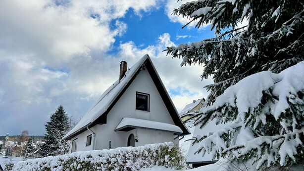 A snow-covered house with a steep roof, surrounded by snow-laden trees and bushes.