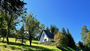A house with a sloped roof, surrounded by lush greenery and a clear blue sky.
