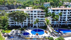 A multi-story hotel with a pool area, including a large central pool and smaller pools, surrounded by palm trees and a lush garden.