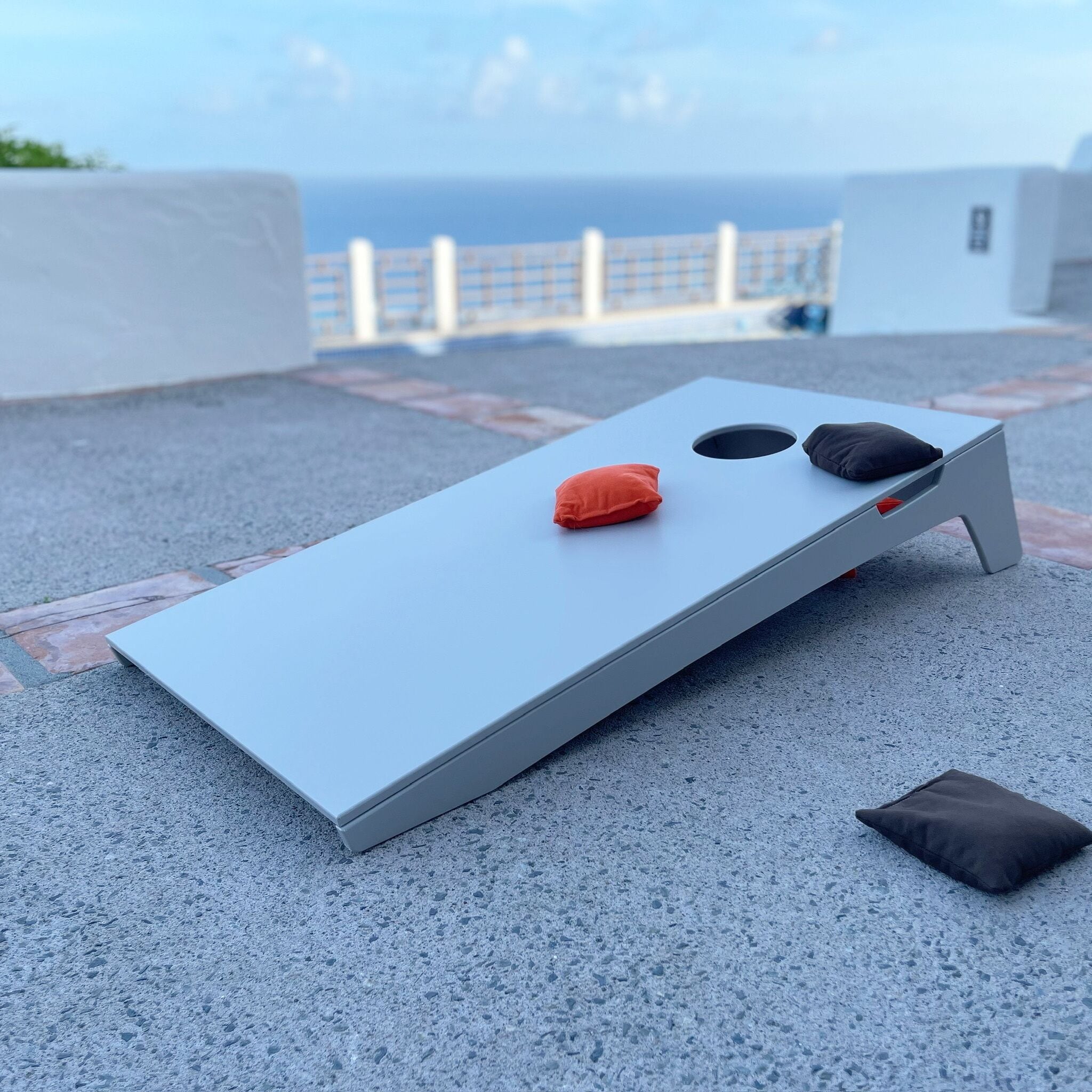 A modern, minimalist cot with a circular cutout, two pillows, and a small cushion on a rooftop terrace with a view of the sea.