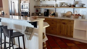 Una cocina con una isla central, taburetes de bar, armarios de madera y estantes con cerámica y vajilla.