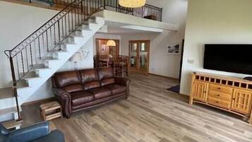 Un vaste espace de vie comprenant un escalier, un canapé en cuir, un pouf et un téléviseur posé sur un meuble en bois.