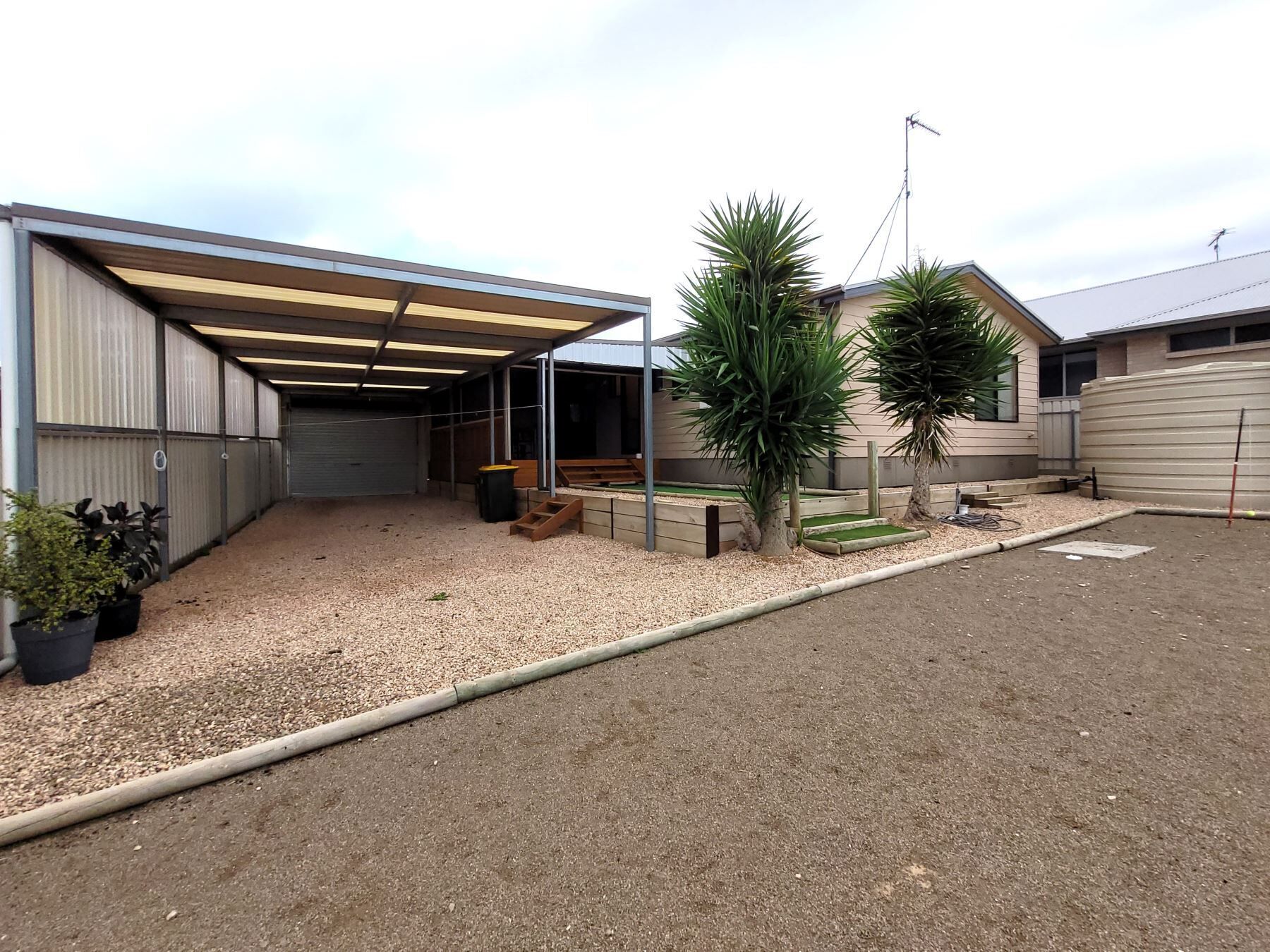 A covered outdoor area with a gravel floor, a small wooden bench, and potted plants.
