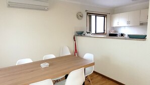 Une cuisine de taille réduite avec une table en bois et des chaises blanches, une horloge au mur et une fenêtre donnant sur un paysage agréable.