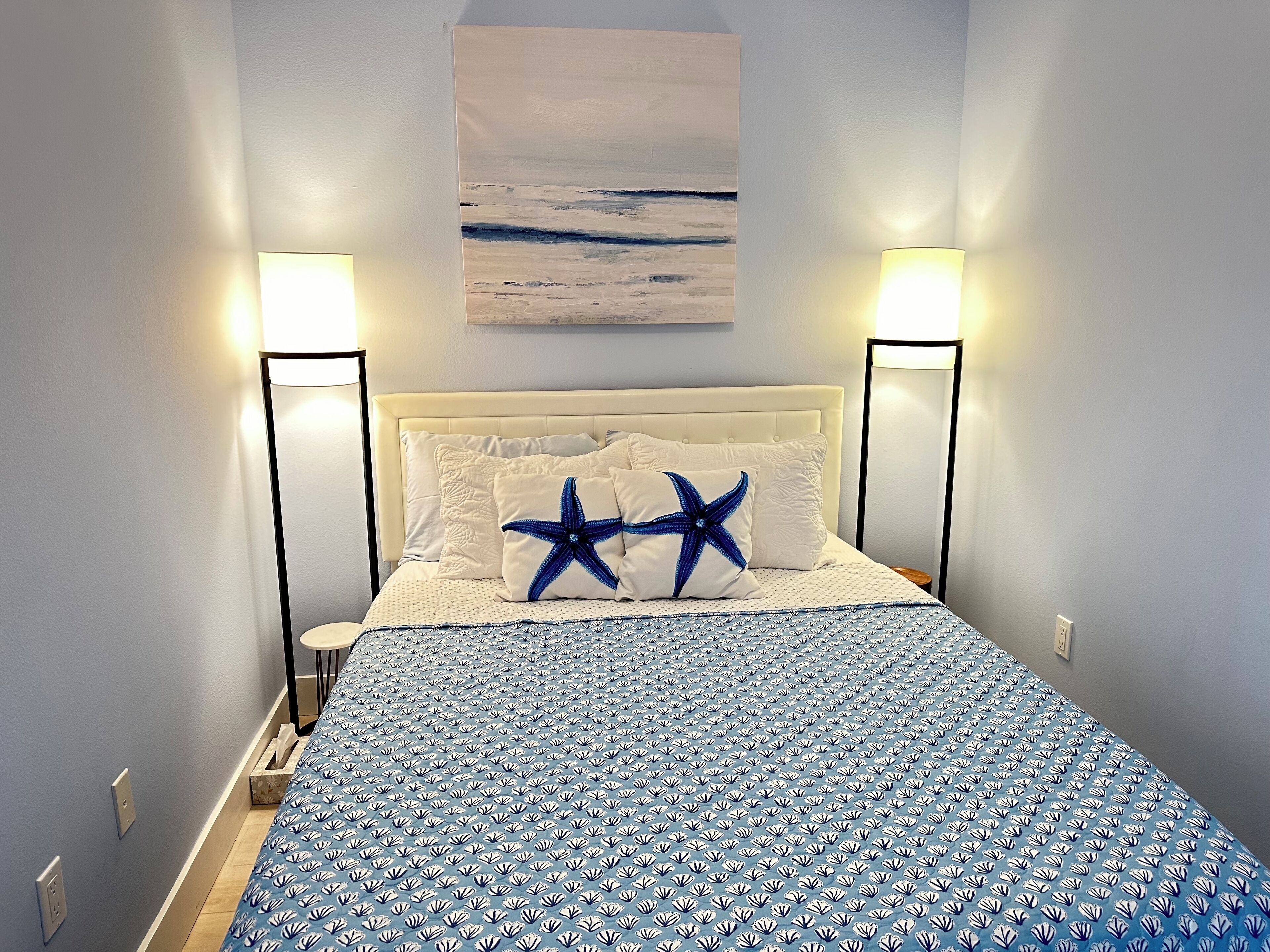 A bedroom with a bed, two bedside lamps, a starfish pillow, and a painting on the wall.