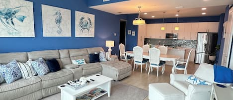 Una sala moderna con un sofá seccional color beige, una mesa de centro blanca y un comedor con sillas blancas.