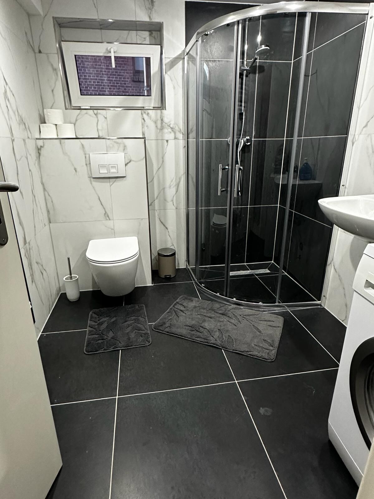 A modern bathroom with a glass shower enclosure, a toilet, a sink, and a small window above the toilet.