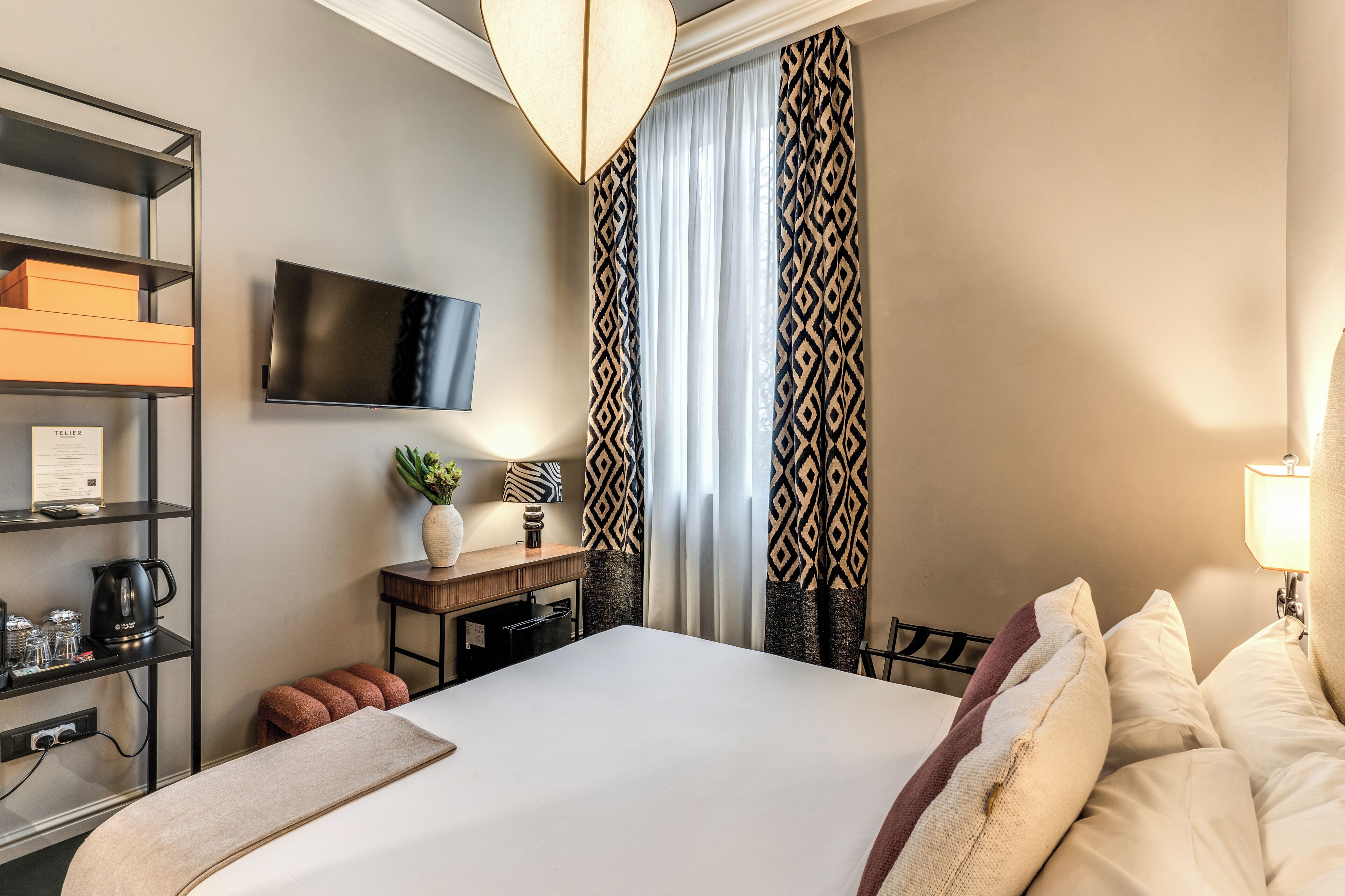 Una habitació d'hotel amb un llit, televisió, escriptori amb làmpada, prestatges amb objectes i cortines amb dibuixos.