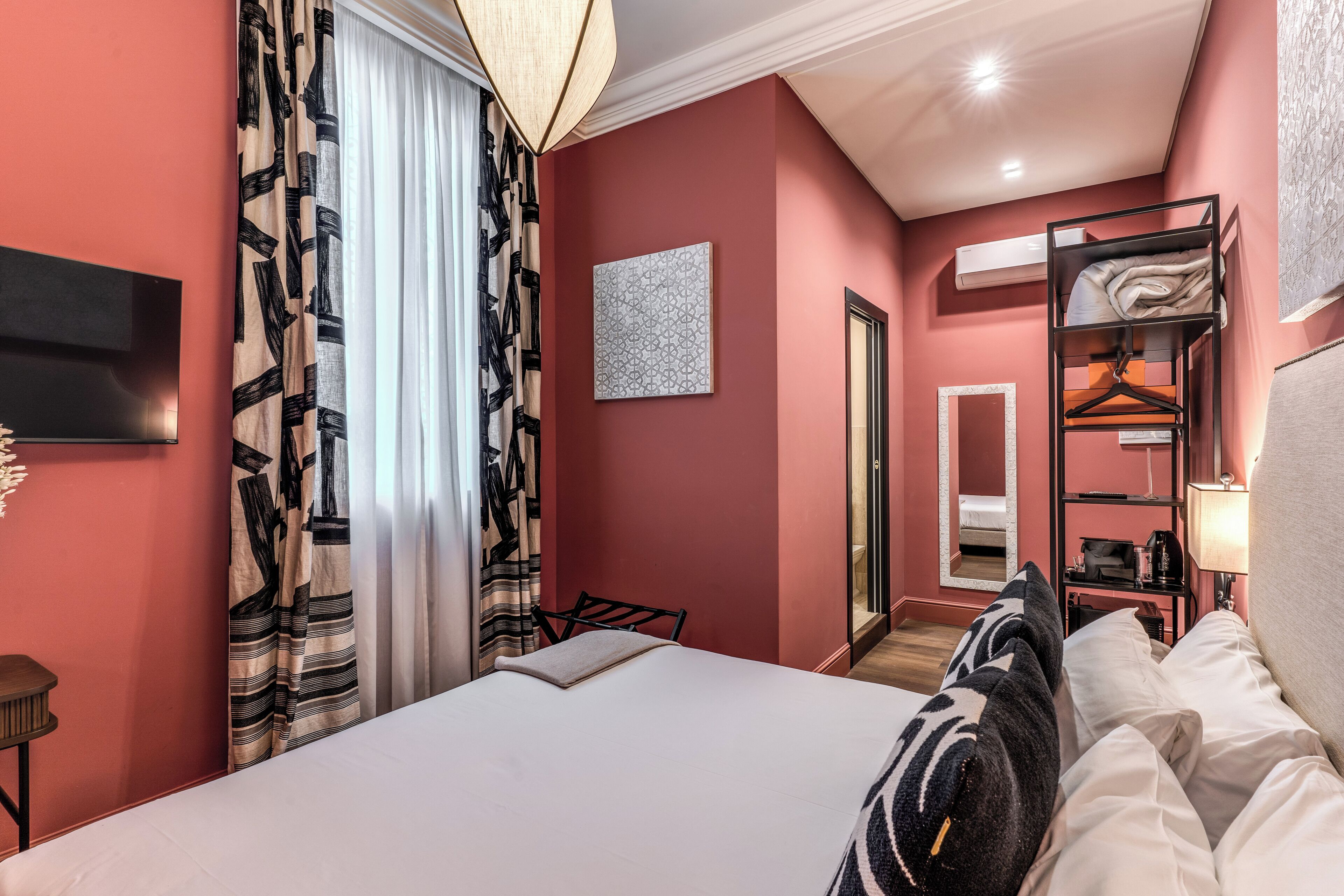 A hotel room with a bed, a bedside table, a hanging wardrobe, a mirror, and a painting on the wall.