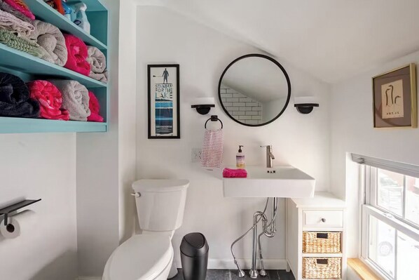 A bathroom with a toilet, sink, and shower. There is a shelf with towels and a framed picture on the wall.