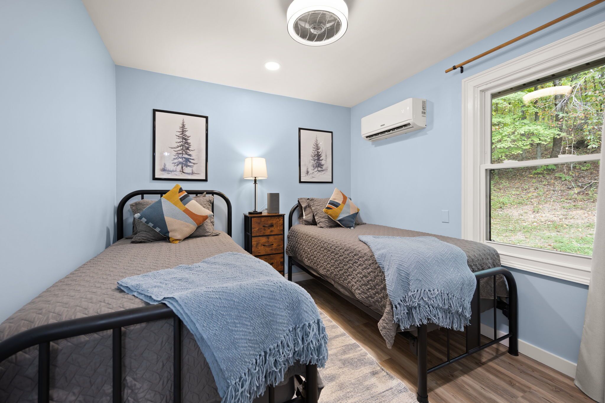 Une chambre avec deux lits, une table de chevet, une lampe et une fenêtre donnant sur des arbres.