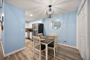 Un coin repas avec une table dressée pour quatre personnes, des chaises blanches, un lustre et une cuisine en arrière-plan.