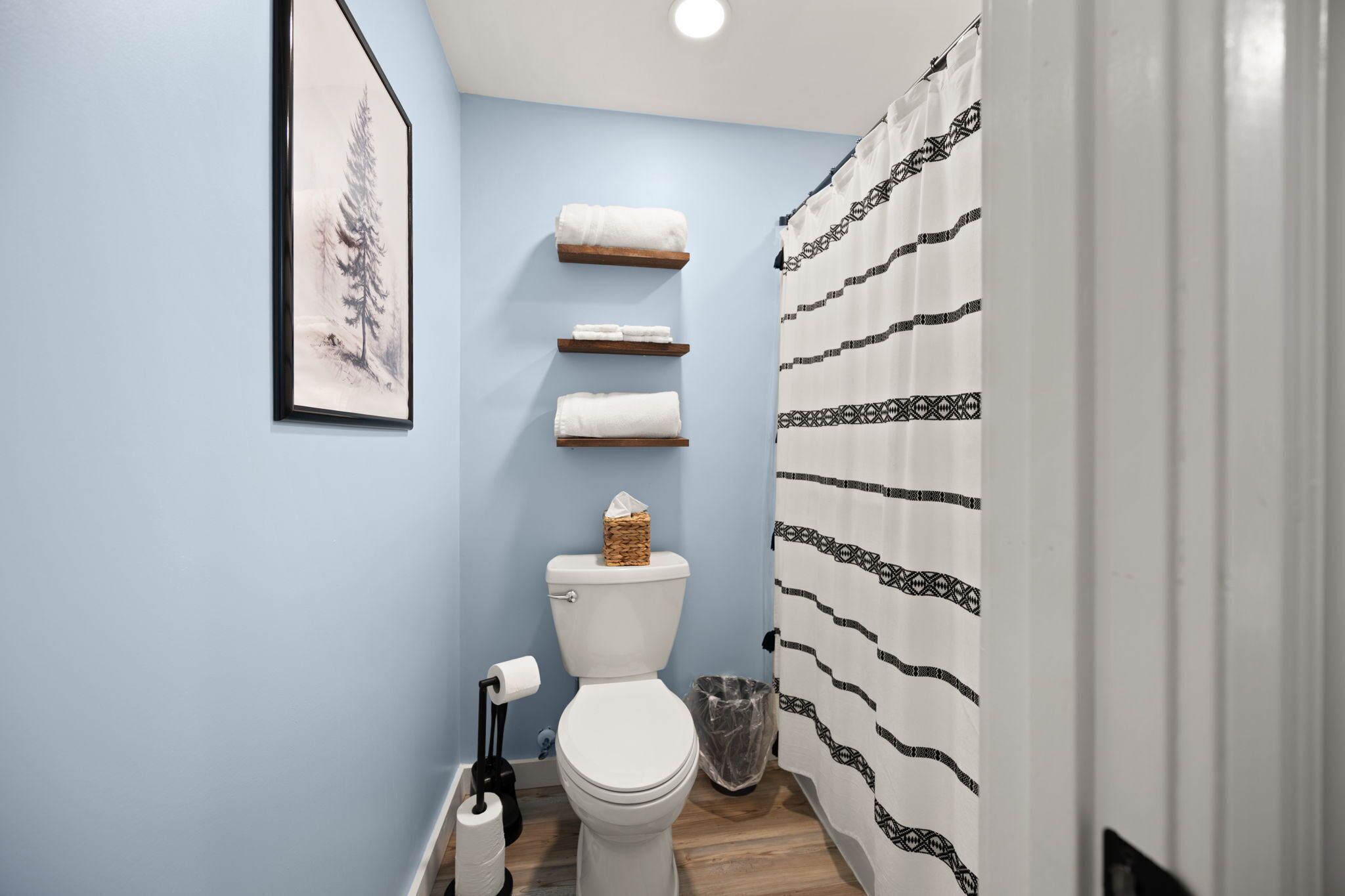 Un baño con inodoro, repisas con toallas, un cuadro en la pared y cortina de ducha estampada.