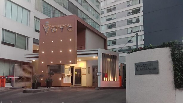A modern hotel entrance with a prominent sign, a parking area, and a tall building in the background. - WTC Suites (Mexico City)