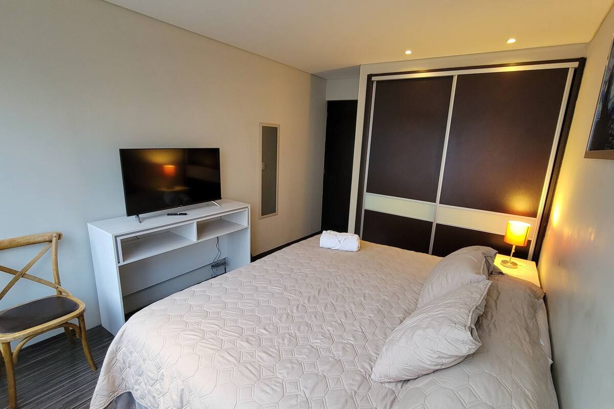 A hotel room with a bed, a TV on a white stand, a chair, and a nightstand with a lamp.