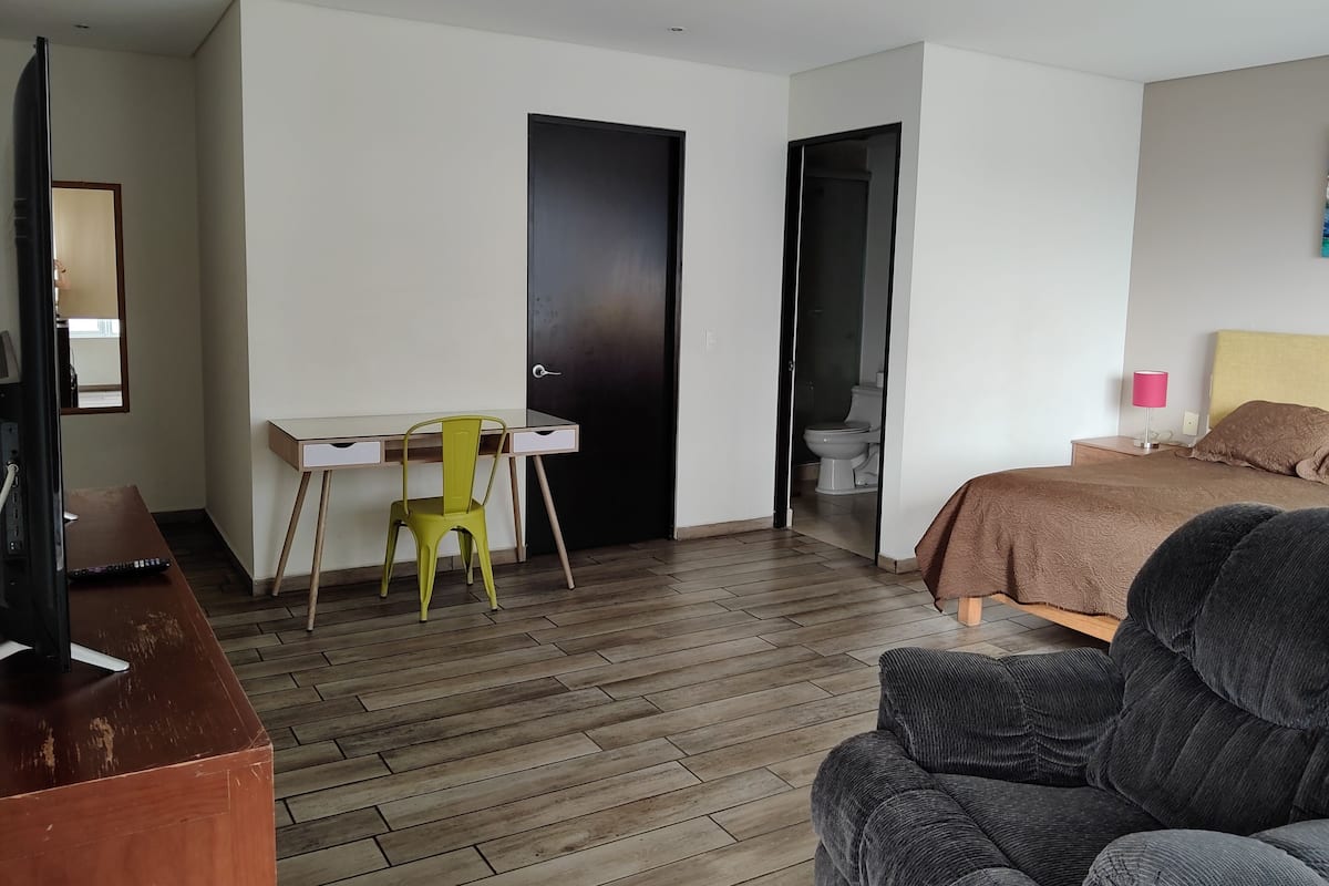 A room with a bed, a desk with a chair, a television, and a bathroom visible through an open door.