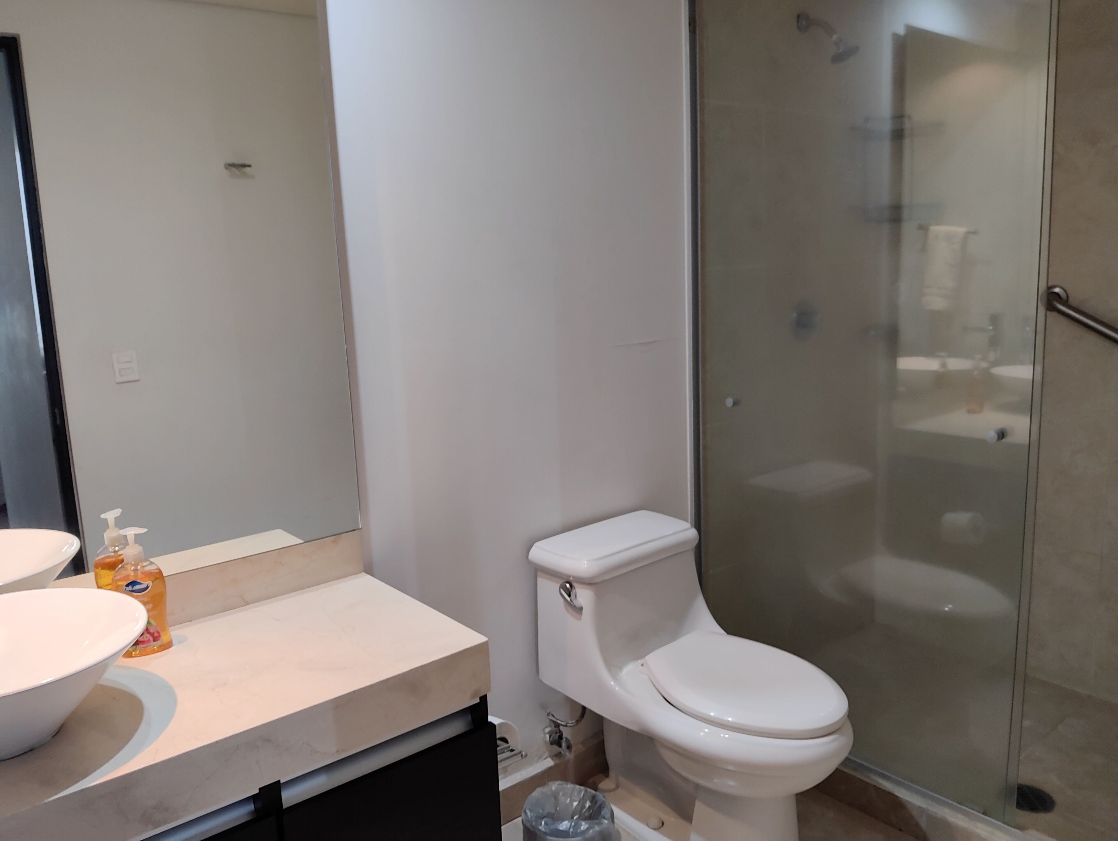 A modern bathroom with a white toilet, a glass-enclosed shower, and a beige countertop with a soap dispenser.