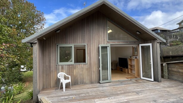A wooden cabin with a porch, a chair, and a sliding door leading to an interior space. - The Guesthouse, Campbells Bay. (Auckland)