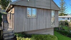 A small wooden cabin with a sloped roof, a front porch, and a small deck. - The Guesthouse, Campbells Bay. (Auckland)
