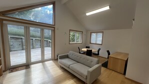 A spacious room with a large sliding glass door leading to an outdoor deck, a grey sofa, a small round table with chairs, and a wooden cabinet. - The Guesthouse, Campbells Bay. (Auckland)