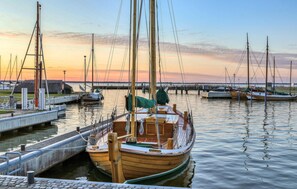 Ein hölzernes Boot liegt in einer Marina festgemacht, mit anderen Booten, einer Mole und einem Sonnenuntergang im Hintergrund.