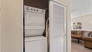 Een wasruimte met een wasmachine en droger, een gesloten kastdeur en een bruine bank in de aangrenzende woonkamer.