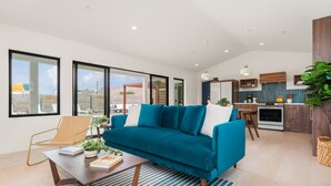 A modern living room with a teal sofa, a wooden coffee table, and a view of the kitchen and outside. - Hoku House - Heated Pool/Spa - Luxury Walkable Oasis in Downtown Joshua Tree (Joshua Tree)