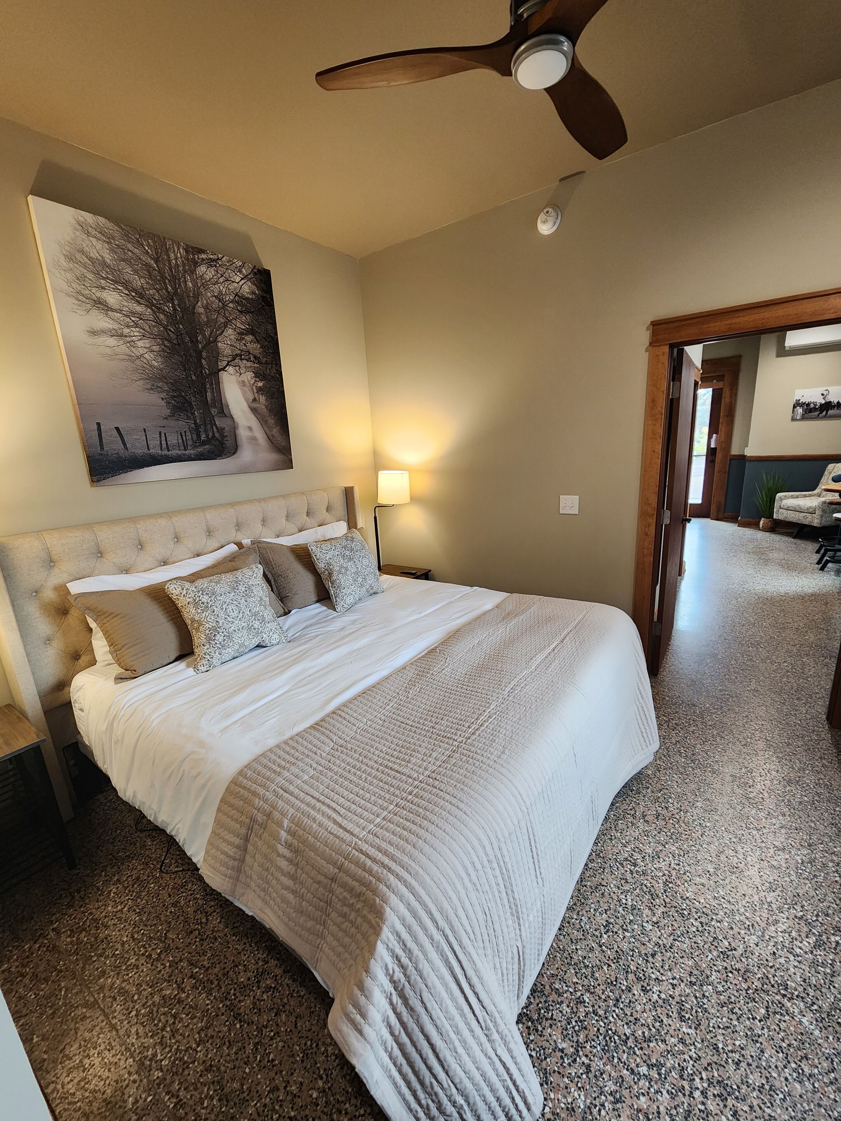 A bedroom with a large bed, a ceiling fan, a nightstand, a lamp, and a painting on the wall.