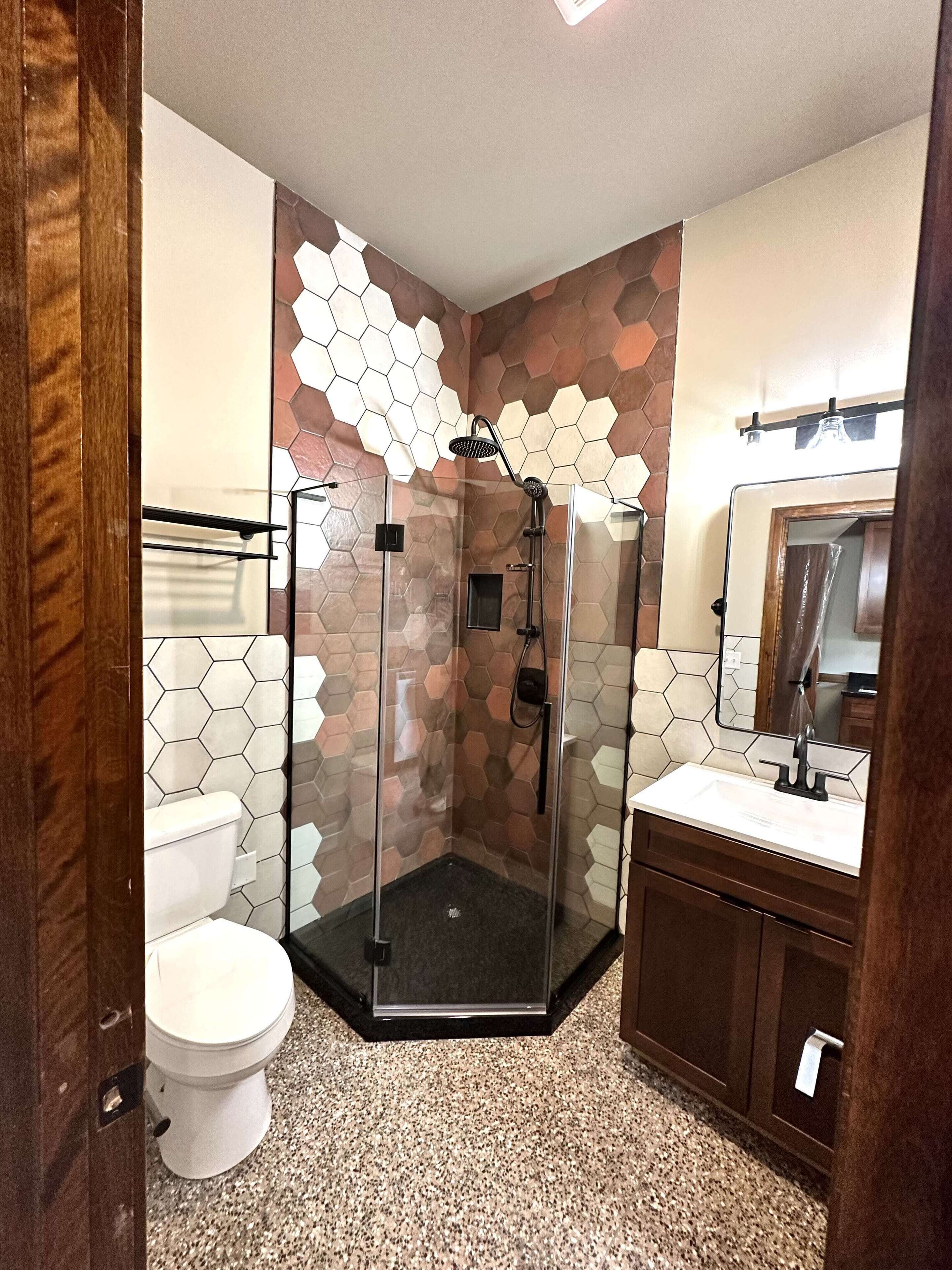 Een badkamer met een wand van hexagontegels, een douche met glazen wand, een wit toilet en een houten wastafelmeubel met spiegel.