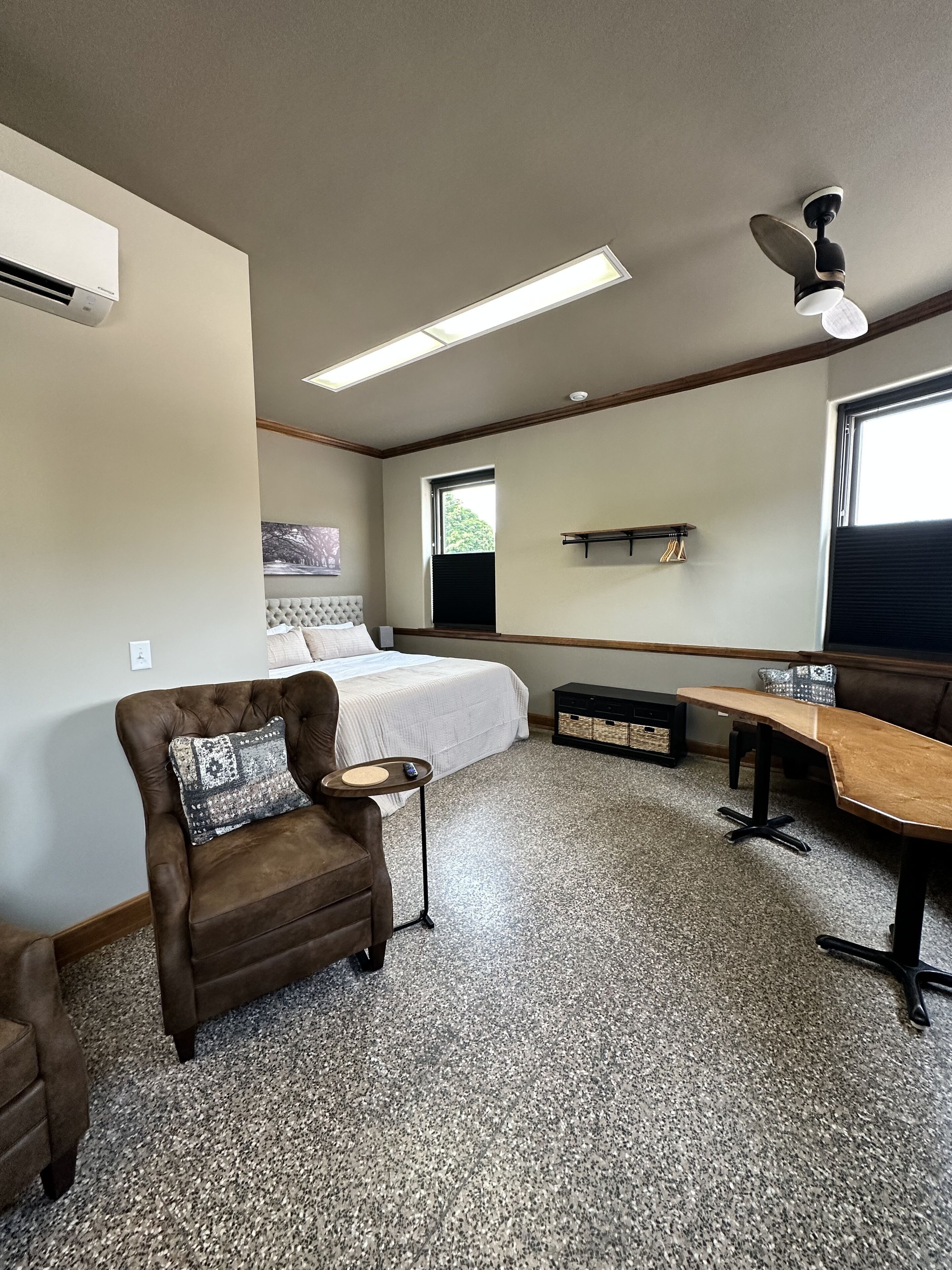 A bedroom with a bed, a brown armchair, a small table, a ceiling fan, and a window.